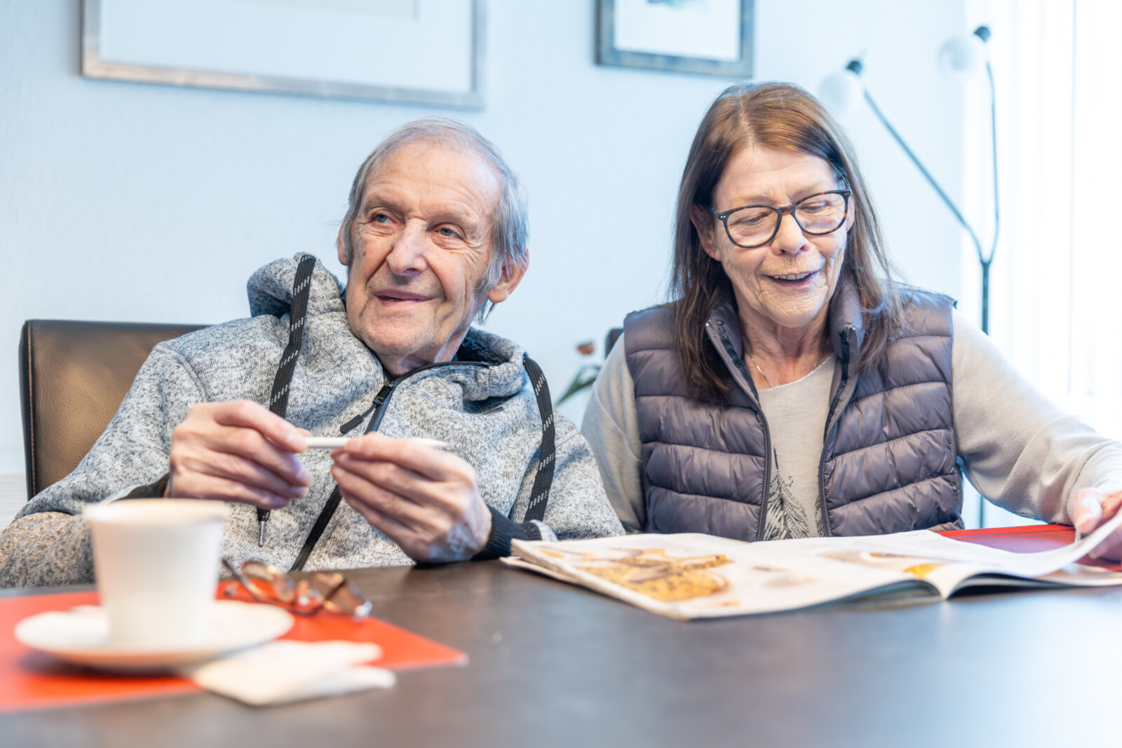 Mann mit grauen Haaren und grauer Kapuzenjacke hält Stift in der Hand, Frau mit braunen Haaren und gesteppter Weste sitzt daneben am Tisch mit aufgeschlagener Zeitschrift und Kaffeetasse