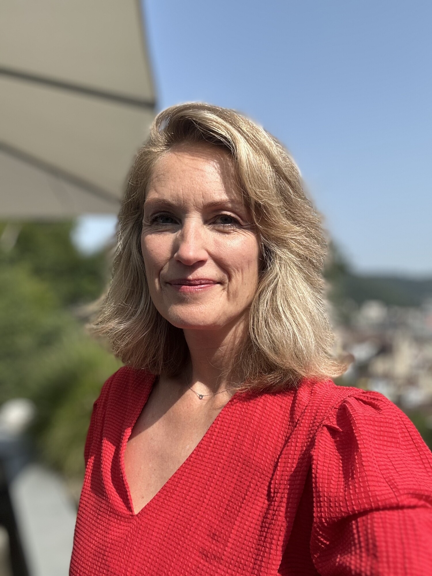 Frau mit schulterlangen blonden Haaren und rotem T-Shirt blickt freundlich in die Kamera, im Hintergrund verschwommen ein Sonnenschirm.