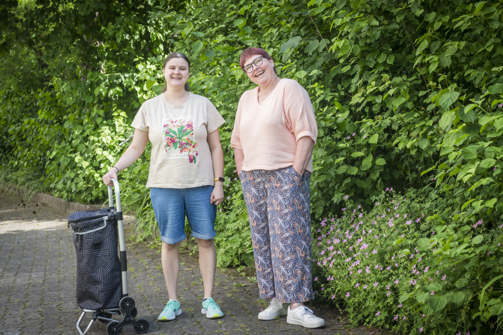 Zwei Frauen stehen nebeneinander auf einem Gehweg, eine mit Einkaufstrolley, beide lässig gekleidet vor einer grünen Hecke mit kleinen rosa Blüten.