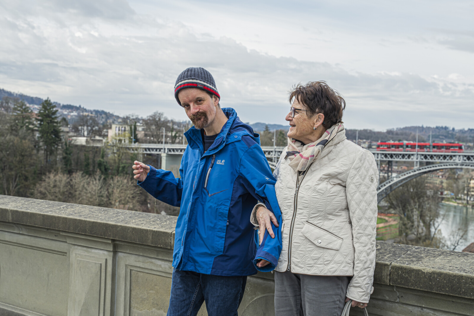 Mann mit blauer Jacke und Mütze hält die Hand einer Frau mit heller Steppjacke und Schal an einer Brüstung mit Fluss und Tram im Hintergrund.
