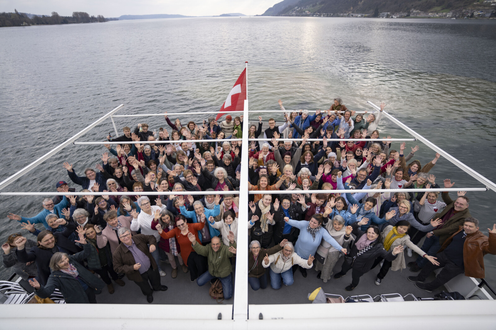 Gruppe von Menschen auf einem Boot vor einem See mit Schweizer Flagge, die in die Kamera winken.