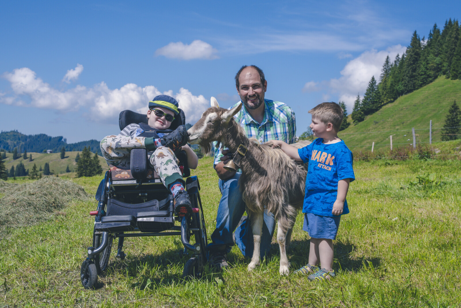 Ein Vater mit seinen zwei Kindern, eines davon im Rollstuhl, stehen auf einer Wiese mit einem Ziegenbock, im Hintergrund Hügel und Wald