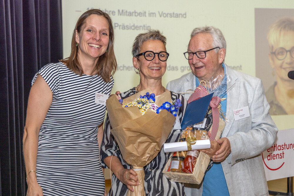 Drei Personen stehen nebeneinander, die mittlere hält einen Blumenstrauss, die rechte einen Geschenkkorb und eine Urkunde, im Hintergrund eine Projektionswand mit Text und einem Porträtfoto.