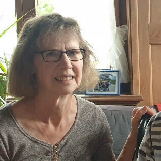 Eine Frau mit Brille und schulterlangem Haar sitzt in einer Wohnstube mit viel Holz und blickt in die Kamera.